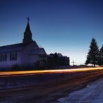 A rural church seen from the road in the early morning just before sunrise, with a streak of light from a vehicle’s headlights running down the road (from a long-exposure photo).