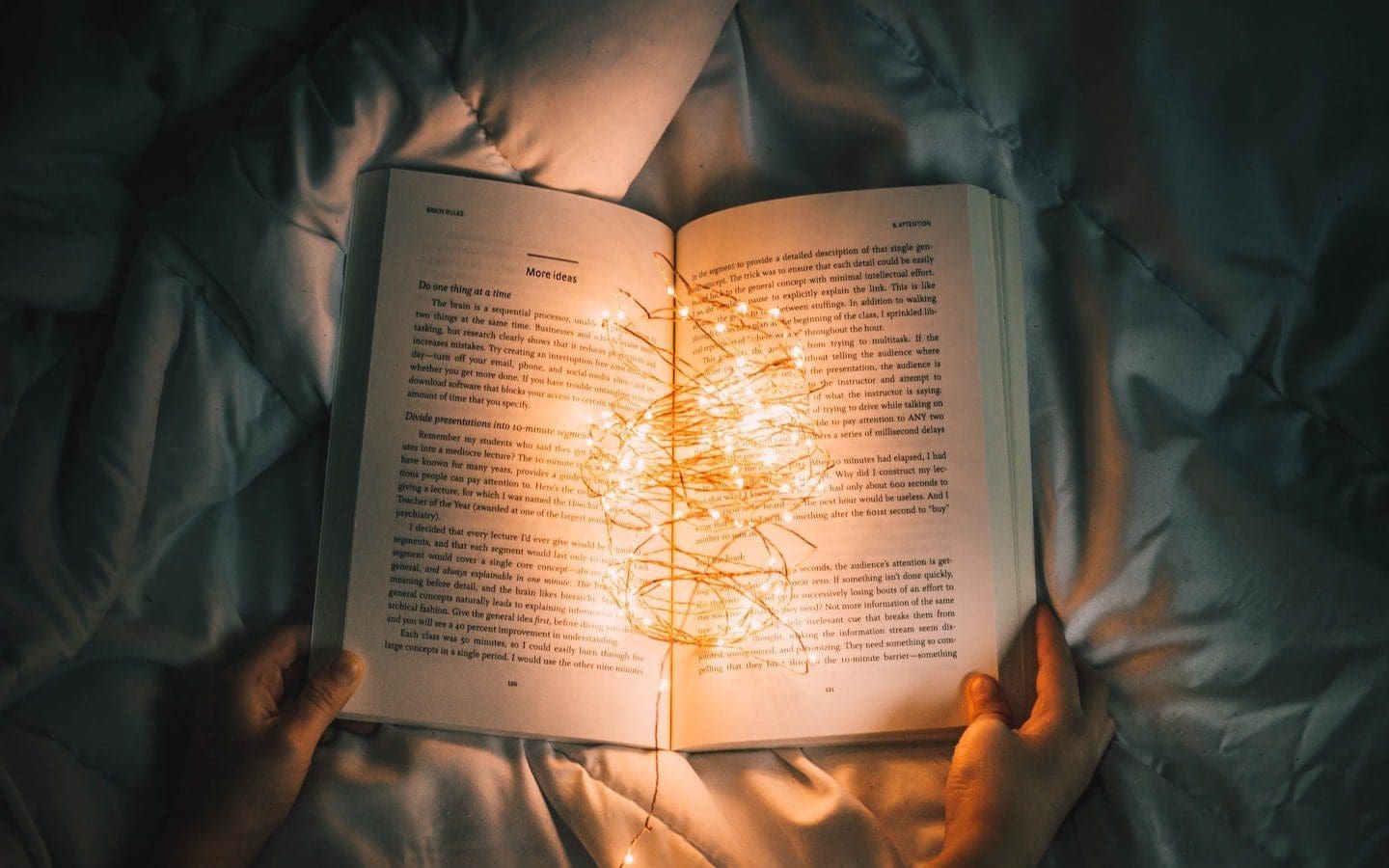 An open book sitting on a bed with a string of lights on top of it illuminating the pages.