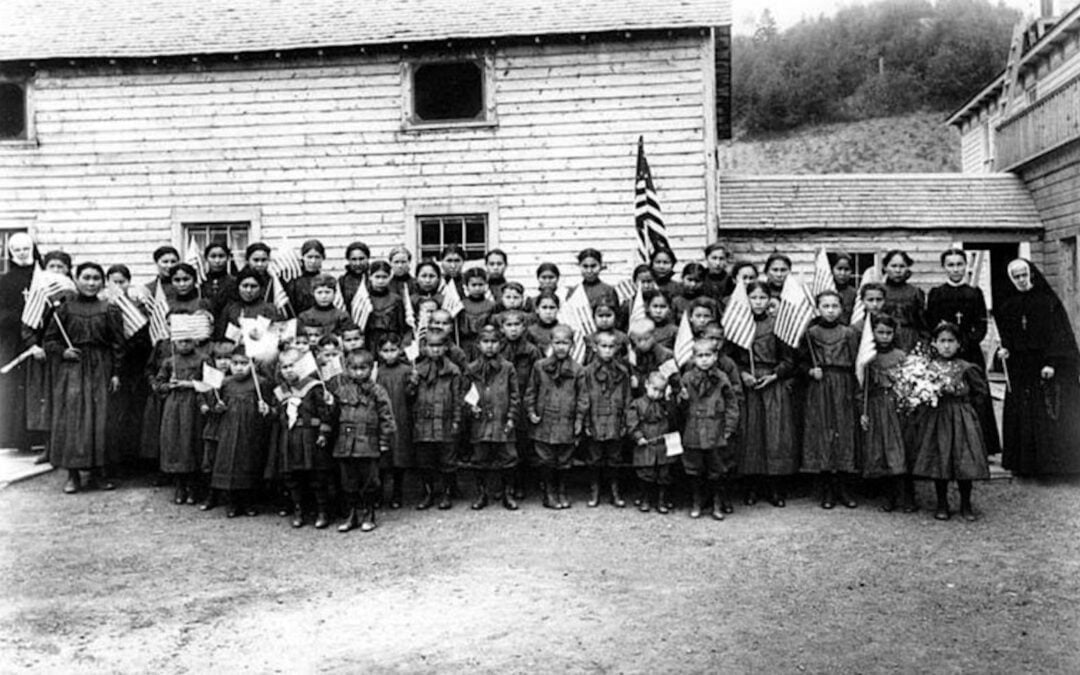 Indigenous children in school uniforms holding American flags in front of a mission school in Holy Cross, Alaska.