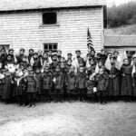 Indigenous children in school uniforms holding American flags in front of a mission school in Holy Cross, Alaska.