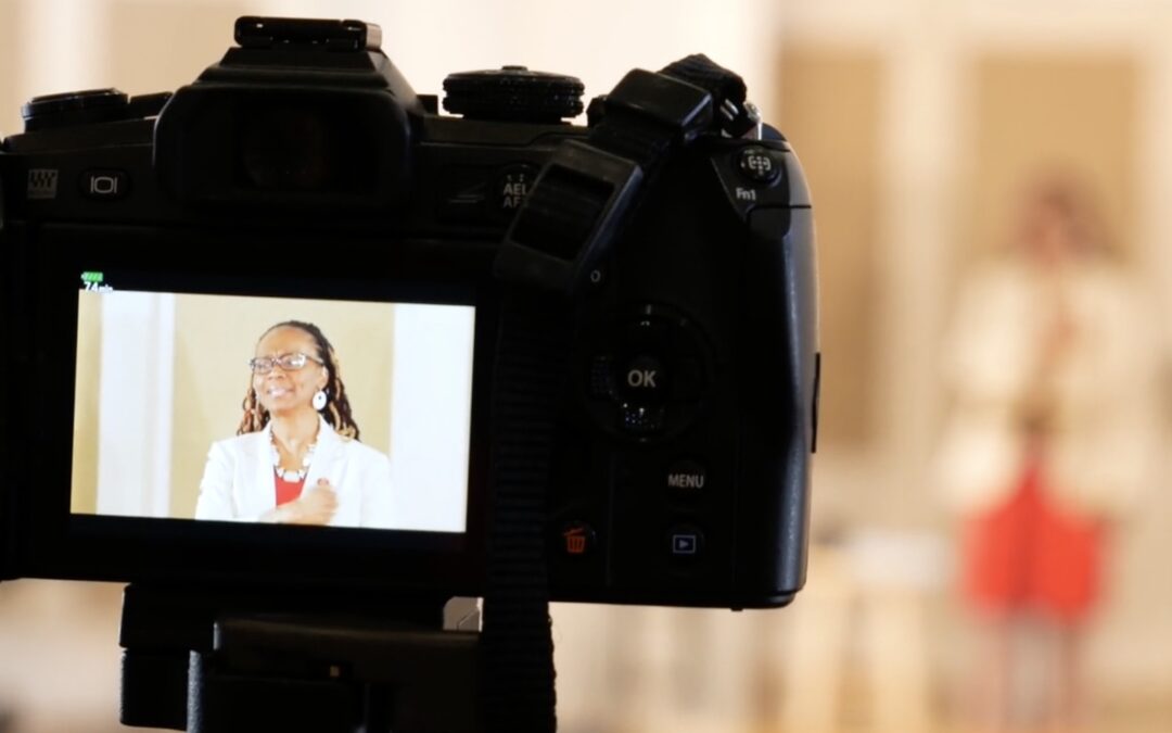 A video camera seen up close with a woman on the camera’s preview screen.