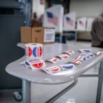 A table with “I voted” stickers on a table in the foreground with people sitting at voting booths casting their ballots in the background.