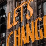 A yellow, metal sign on the sign of a brick building that says, “Let’s Change.”