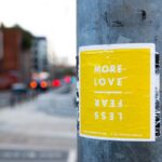 A yellow sign on a metal light pole that says, in white lettering, “More Love” and then below it and upside down, “Less Fear.”