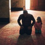 A father and daughter kneeling side by side on a rub praying in a mosque.