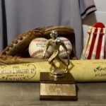 Various baseball items – trophy, wooden bat, glove, balls – sitting on a wooden table, with a gray jersey hanging up in the background.