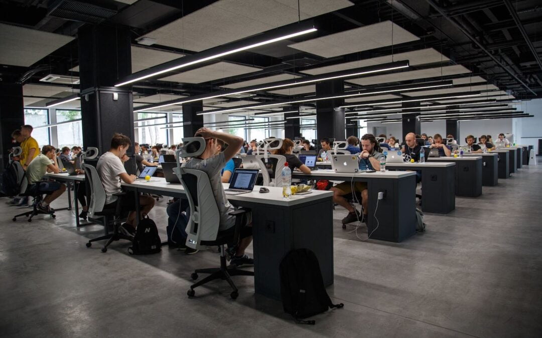 A large room in an office building with people sitting at long tables working at computers.