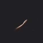 A person’s hand and forearm seen in a small amount of light reaching out in a completely dark space.
