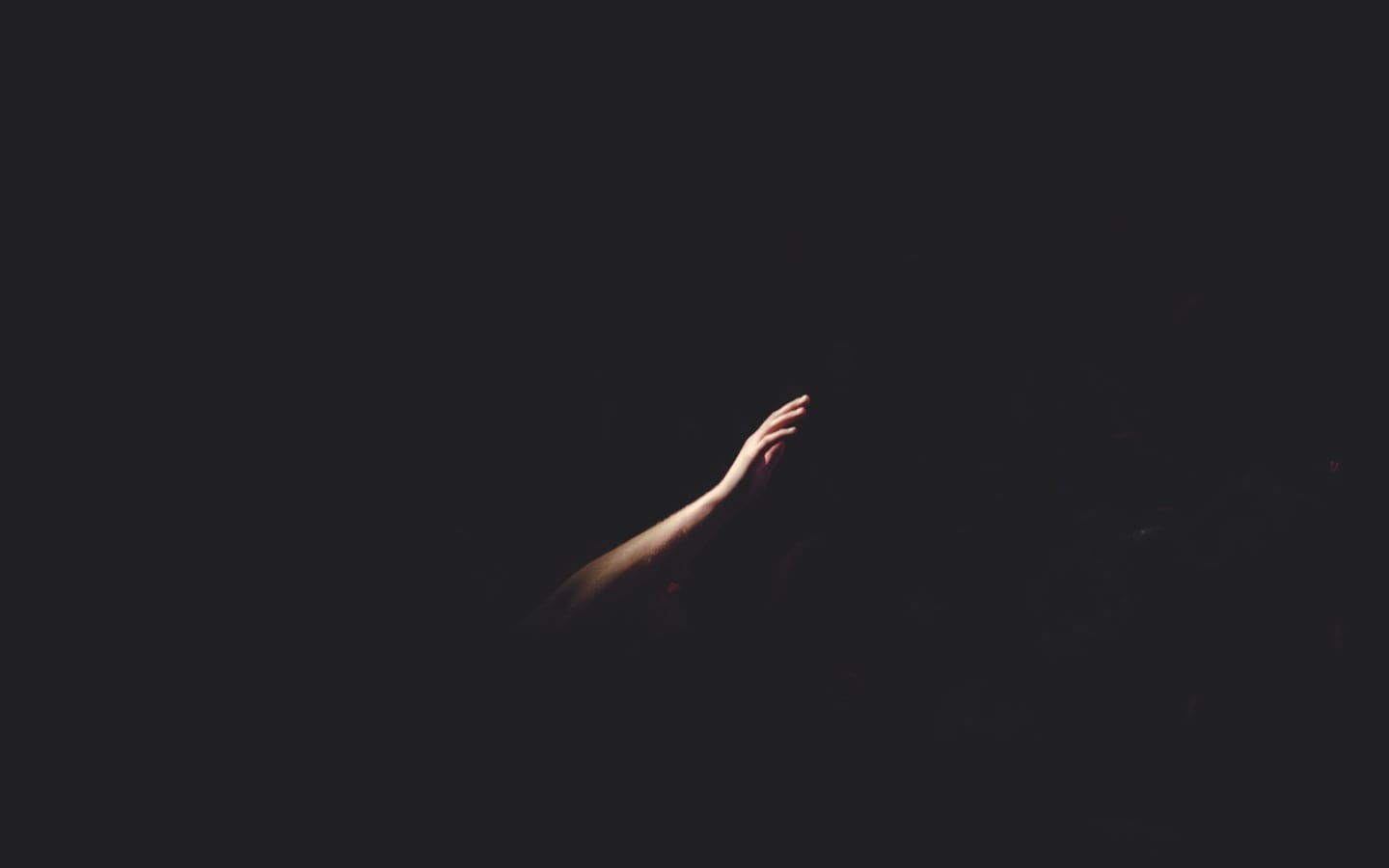 A person’s hand and forearm seen in a small amount of light reaching out in a completely dark space.