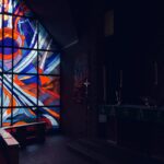 The interior of a church worship space with the room dark except for light shining through a stained-glass window.