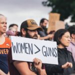 A man holding a sign that say, “Women > Guns” in a group of people standing together outside.