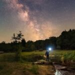 A person standing outside next to a stream holding a light while looking up at the stars.
