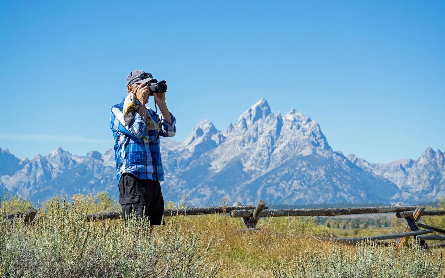 A man standing in a field taking a photo with mountains in the background.