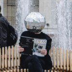 A person with a disco ball on their head holding a magazine while seated on a bench.