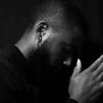 An African American with his hands pressed together and his head bowed against a black backdrop.