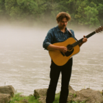 Zach Russell standing alongside a river with a guitar in hand.