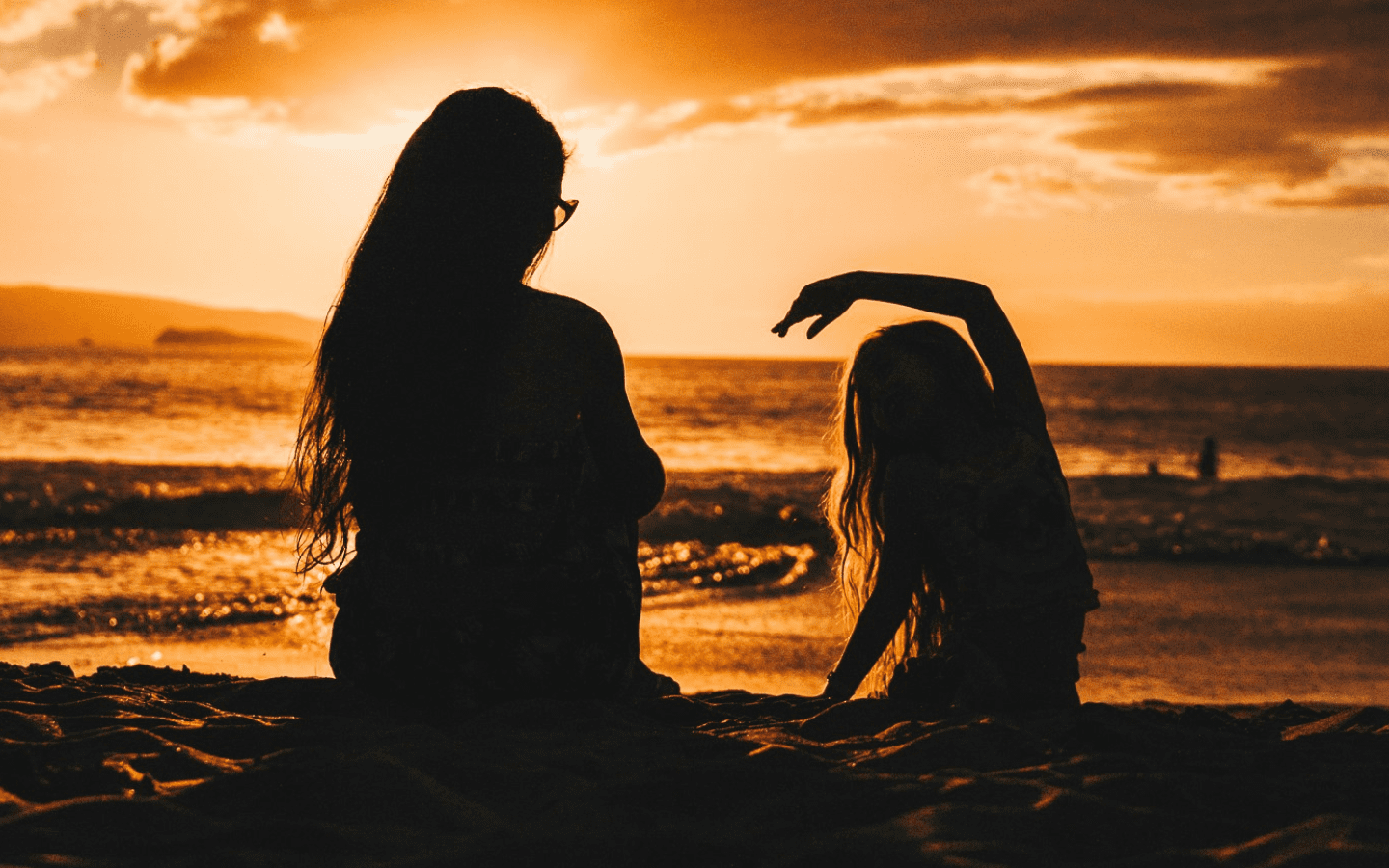 A mother with her young child sitting on a beach at sunset.