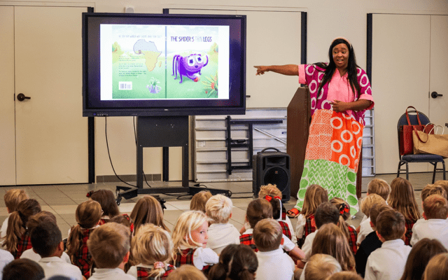 A woman standing in front of a television screen pointing to a book and children looking on.||