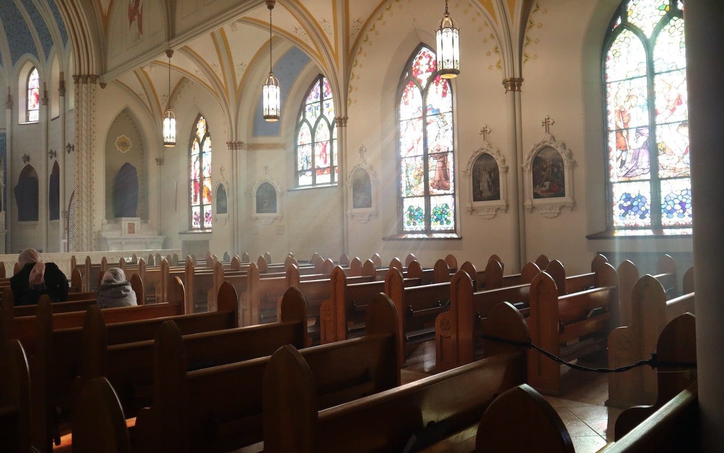 The interior of a church with a few people in the pews and sunlight shining in through stained glass windows.