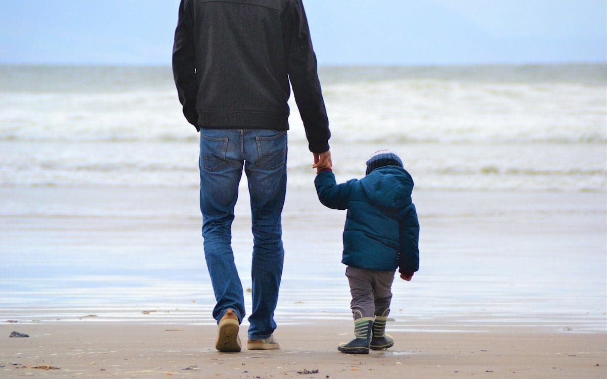 A dad walking on the beach holding his young child's hand.|Dad carrying son on his shoulders