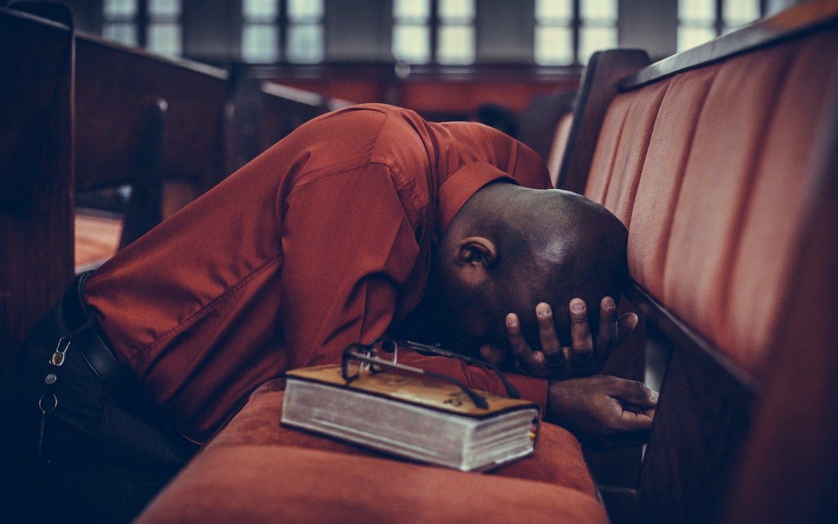 A man on his knees in a church pew with his head in his hands and a Bible sitting next to him.