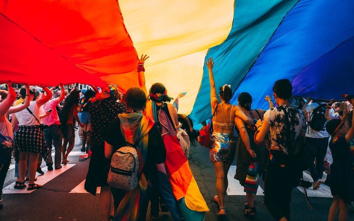 A rainbow flag in the air with several people standing underneath.
