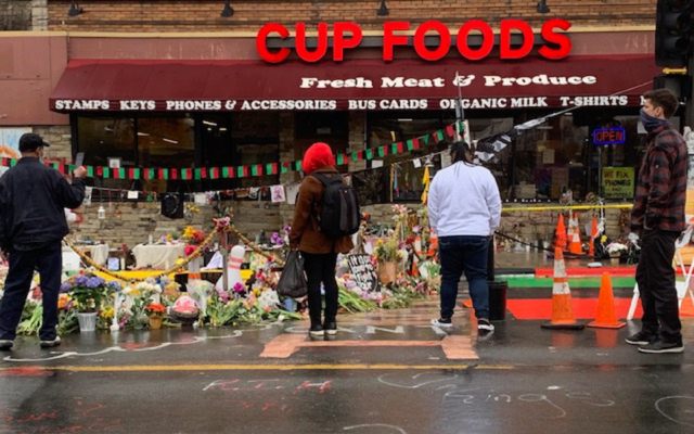 People standing in front of a neighborhood store that doubles as a memorial for a man murdered just outside its doors.